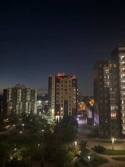 This is a medium-shot, vertical-orientation photograph taken at night, capturing a view of apartment buildings and a park-like area in Ligachevo, Russia. The sky is a deep navy blue, with a few faint stars visible.
In the foreground, a well-lit park area features winding pathways, green grass, and numerous streetlights illuminating the scene. Trees with dense foliage are scattered throughout the park, casting shadows.
Dominating the midground are three tall, multi-story apartment buildings. The building on the far left is lighter in color with green and beige sections, featuring many illuminated windows. The central building is taller and darker, with a mix of brown and beige facades, also showing numerous lit windows. The building on the far right is the tallest and darkest, with a predominantly brown facade and very bright, illuminated windows. The tops of the central and right buildings have red lights.
In the background, more apartment buildings and some industrial structures are visible in the distance, with lights from various sources indicating an urban or suburban environment. There are also power lines visible in the middle distance. The overall impression is a view of a residential area at night, with an emphasis on the illuminated buildings and the park. No people are discernible in the image.