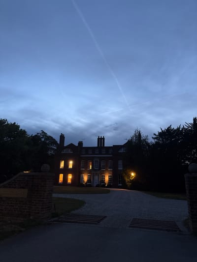 A nighttime, low-angle, full shot captures a large, traditional brick house situated in the city of Chelmsford, United Kingdom. The sky above is a deep twilight blue with wispy clouds and two faint contrails visible. The house features multiple stories, brick chimneys, and numerous windows, many of which are illuminated with a warm, inviting light. Lush, dark green trees frame the property on either side. In the foreground, a gravel driveway leads up to the house, bordered by dark asphalt. To the left, a brick gatepost stands with a weathered sign that reads "Swaffham Place". A metal cattle grid is installed across the driveway. The scene suggests a serene and established residential setting at dusk.