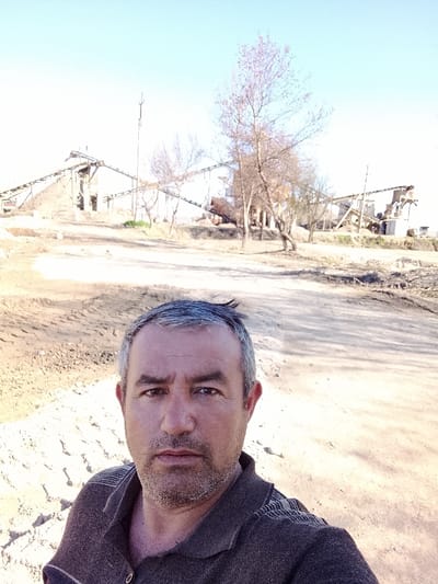 An adult male is prominently featured in the foreground of a daylight selfie, positioned in Yangi Chinoz, Uzbekistan. He has graying dark hair, a short beard, and a neutral expression, looking directly into the camera. He is wearing a dark brown collared shirt with horizontal stripe patterns on the shoulders. The background reveals an industrial or construction environment characterized by light brown, uneven ground with visible earth disturbances. Multiple industrial structures dominate the mid-ground, including several interconnected conveyor belt systems supported by metal frameworks, leading to and from large aggregate piles and processing machinery. A tall, slender pole stands vertically near the center of the background. Scattered leafless deciduous trees are present throughout the scene. The sky is clear and light blue, indicating fair weather conditions.