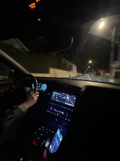 A dark interior car view shows a person driving at night, holding a phone displaying a map while presumably following directions on the car's central navigation screen. The road ahead, illuminated by sparse streetlights, stretches into the darkness. The passenger airbag light is visible. The steering wheel is in the driver's hands, which are illuminated slightly by the car's interior lights.