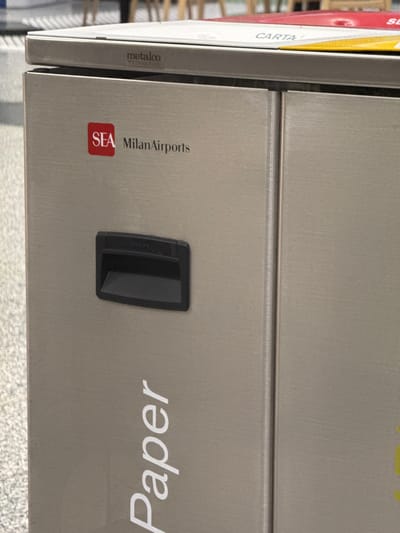 This is a close-up, vertically oriented photograph of a modern waste bin, likely made of brushed metal. The bin has a sleek, minimalist design with a horizontal seam dividing its upper and lower sections. On the left side of the bin, a white label with the text "CARTA" is partially visible, indicating that this section is designated for paper waste. Below this, the name "metalco" is printed in a smaller font.
The right side of the bin features the word "paper" printed in large, white, stylized lettering, also indicating its purpose. Below the "paper" text, there is a red square logo with white lettering that reads "SEA MilanAirports," suggesting the bin is located within Milan Malpensa Airport or Linate Airport, both operated by SEA. A black, rectangular handle or opening mechanism is positioned in the center of the lower section of the bin.
The background is blurred but appears to be an indoor environment, possibly an airport terminal, with hints of shelving units and other objects. The lighting is even, suggesting an interior setting, and there are no indications of weather or time of day. The overall impression is one of functionality and modern design, typical of public spaces.