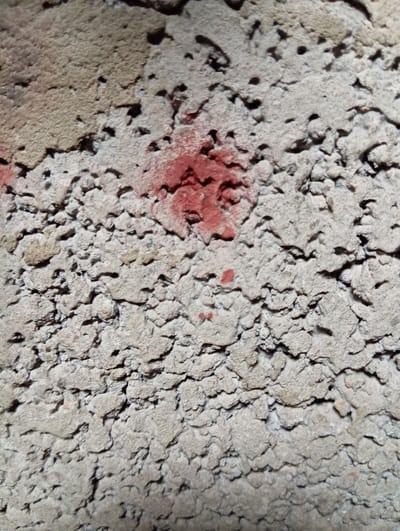 The image shows a close-up view of a rough, porous concrete surface with red spray paint marks. The concrete has an uneven texture with many small holes and crevices. There are two visible red paint marks. One is a larger, more amorphous splotch located slightly above the center of the image. Below and to the left of this larger mark, there are smaller, scattered red paint dots. The lighting suggests the surface is outdoors, possibly a wall or a construction material. The location in Hlynytsi, Ukraine, is noted, but no specific environmental or urban context beyond the concrete surface itself is discernible from the image.