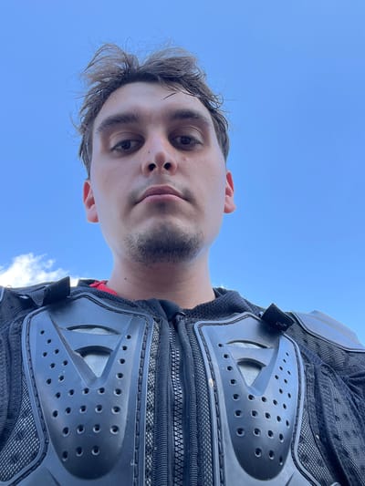 This is a close-up, low-angle selfie of a young man wearing protective motocross gear. The man has short, dark, messy hair and is looking directly at the camera. He has dark eyebrows, brown eyes, and a faint shadow of stubble on his chin and upper lip. He is wearing a black motocross chest protector with multiple holes and a V-shaped design on the front, and a red shirt is visible underneath. The background is a clear blue sky with a few wispy white clouds on the left side. The image was taken outdoors. The location is not specified in the image or its metadata.