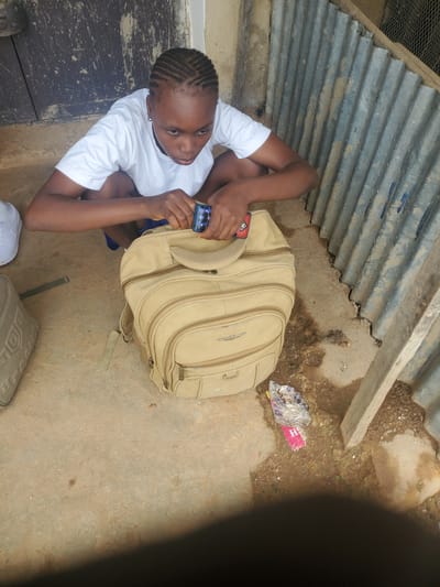 A young Black person with braided hair is crouched down next to a tan backpack. They are wearing a white t-shirt and dark shorts. Their hands are in the process of opening a small, dark blue object, possibly a phone or a small electronic device. The backpack is large and appears to be made of canvas, with multiple pockets and zippers. To the left of the backpack is another bag, a lighter tan color, with a patterned design. The ground is a dusty, sandy surface. Behind the person and backpack is a corrugated metal wall, showing signs of rust and dirt. To the left of the metal wall is a wooden door with peeling paint. The overall environment suggests an outdoor setting, possibly a yard or alleyway, in a somewhat weathered and utilitarian area. The presence of trash, including a pink item with the letter "H" and a clear plastic bag filled with white objects, further indicates a less-than-pristine location.
