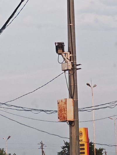 The image captures a traffic camera mounted on a utility pole in Nizhnyaya Elyuzan, Russia. The camera is positioned at the top of the pole, facing forward. Below the camera, a rusty metal box is attached to the pole. Electrical wires are strung across the scene, connecting to the utility pole and extending into the distance. In the background, a streetlamp and a gas station sign with prices displayed are visible. The sky is overcast, suggesting a cloudy day.