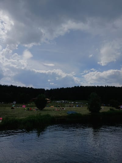 The image captures a daytime scene at a lakeside recreational area, identified as being in the city of Revda, Russia. The foreground features a body of water with ripples on its surface. The middle ground shows a grassy bank dotted with people sunbathing, sitting on chairs, and relaxing under umbrellas and tents. Various objects like beach balls, bags, and a blue kayak are visible on the grass. Several cars are parked in the background, beyond the grassy area, adjacent to a dense line of dark green trees that forms the backdrop. The sky is partially cloudy, with a mix of dark and light gray clouds against a pale blue sky. A few buildings, possibly small structures or shelters, are partially obscured by the trees.