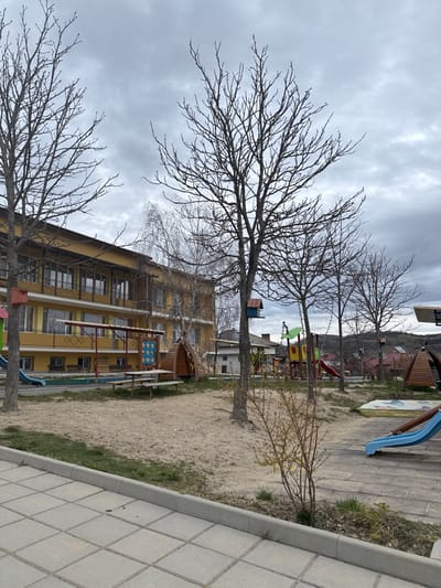 The scene depicts an unoccupied outdoor playground in Breznitsa, Bulgaria, viewed under an overcast sky. The foreground consists of grey square paving stones, transitioning into a sandy, light brown ground with sporadic patches of sparse green grass. To the left, a multi-story building with a yellow facade features numerous windows, balconies with yellow railings, and protective awnings above lower-level windows. Several bare deciduous trees, indicative of late winter or early spring, are positioned throughout the sandy area; some feature attached birdhouses. Playground equipment includes, from left to right: a blue slide, a wooden picnic table with benches, a vertical activity panel with colorful blue, red, and yellow elements, and multiple wooden A-frame structures. Further right, a larger multi-colored play structure (green, yellow, red) with an attached red slide is visible, alongside another blue slide and additional small wooden hut-like structures. A small, multi-branched shrub with developing yellowish leaves stands in the right foreground. Residential buildings and distant hills are visible on the horizon to the right. No individuals are present in the scene.