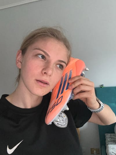 A young adult female is depicted indoors, holding a bright orange and blue Adidas X Speedflow soccer cleat to her right ear, as if listening to it. Her light-colored hair is pulled back, and she has fair skin with faint freckles. Her gaze is directed upward and slightly to her left, with her mouth slightly open. She wears a black short-sleeved t-shirt featuring a white Nike swoosh logo on the left chest and a circular white patch with a soccer ball and Cyrillic text, "ФУТБОЛЬНАЯ МЕЧТА" (Football Dream), on the right chest. A white smartwatch with a perforated band is visible on her left wrist. The background consists of a plain, light-colored wall, with a portion of a teal-colored upholstered object visible on the right. The image does not contain specific visual information to determine its location within Roccavione, Italy.