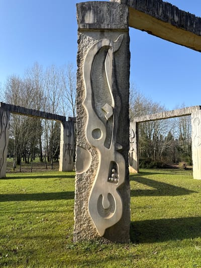 The image depicts an outdoor sculpture installation in a park-like setting within the city of Mealhada, Portugal. In the foreground and central frame stands a weathered concrete pillar, roughly textured, featuring a large, deeply carved abstract design composed of curvilinear and geometric shapes. Multiple similar concrete structures, some serving as vertical pillars with unique carvings and others forming horizontal lintels, are visible in the midground, creating a series of archways or gates. One pillar in the left midground features a carving depicting two stylized humanoid figures beneath a crescent moon. Another pillar to the far right displays a circular, spiral-like motif. The ground is covered in vibrant green grass with areas of sparse vegetation and shadows cast from the structures. In the background, a dense stand of deciduous trees with bare branches extends, indicative of a winter or early spring season. The sky above is clear and bright blue. No individuals are present in the scene. The lighting suggests direct sunlight from the left, creating distinct shadows across the grass.