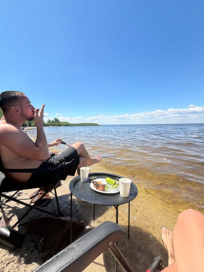 A man sits in a camping chair on a sandy beach in Lebedivka, Ukraine, with the Black Sea stretching out before him under a clear blue sky. He is shirtless and wearing black shorts, with his left arm resting on the armrest of his chair and his right hand bringing a cigarette to his lips. A small black circular table sits beside him, holding a plate of food and two white paper cups. The water laps at the shore, partially submerging the legs of his chair and the table. In the foreground, a portion of another person's legs and feet is visible, suggesting they are also seated in a camping chair. The scene depicts a relaxed outdoor meal by the sea.