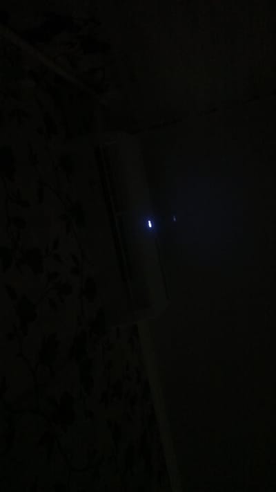 The image depicts a dimly lit interior space. On the left, a wall is covered in dark, floral-patterned wallpaper. To the right, a white, rectangular object with a glowing blue light on its surface is visible. This object appears to be an electronic device, possibly a router or a modem, positioned against a darker background. The overall ambiance is dark, with minimal illumination. No people or specific city details are discernible from the image.