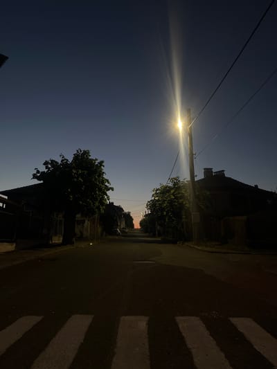 The image depicts a street scene in Gramatikovo, Bulgaria, during twilight. A prominent street light shines brightly, casting a lens flare that streaks across the dark blue sky. Power lines stretch horizontally across the frame, supported by a utility pole. To the right of the street light, silhouetted buildings and trees line the street, with a faint glow of distant lights visible in the background. A pedestrian crossing is visible on the asphalt road, marked by white stripes. The overall mood is serene, with the transition from day to night creating a quiet atmosphere. The time of day is evident from the fading light in the sky and the illumination from the street light. No people are visible in the image.