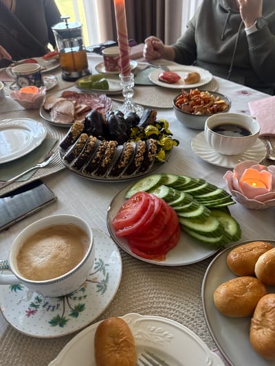The media file captures an inviting dining table set for a meal in an indoor setting, likely a home in Saurieši, Latvia. Several people are partially visible, with one person at the top left wearing a green hooded sweatshirt and a white smartwatch on their wrist.
The table is covered with a light-colored tablecloth and features a variety of food and beverages. On display are plates of sliced cucumbers and tomatoes, small golden-brown bread rolls, and an assortment of chocolate desserts, some topped with crushed nuts and others appearing plain or dark-chocolate coated. Also present are cold cuts, possibly ham and salami, arranged on a plate, and a bowl of small, savory snacks or croutons. Several cups of coffee are visible, some black and some with foamed milk, along with a French press, suggesting coffee is a central part of the meal.
The table is adorned with decorative elements, including a pink rose-shaped candle holder with a lit candle, and another candle in a patterned holder. White plates, some with intricate patterns, and various saucers are arranged, alongside cutlery. A smartphone rests on the table. Small yellow foil-wrapped candies are also visible, with the text "ШОКОЛАДНЫЕ" (Shokoladnye - meaning Chocolate) and what appears to be "JAFFARE" or a similar brand name partially readable on their wrappers.
The atmosphere suggests a relaxed, cozy gathering, possibly a breakfast or brunch, given the combination of coffee, bread, and savory/sweet items. Natural light appears to be present, visible through a window reflection, indicating a daytime meal despite the lit candles which add to the decorative ambiance.