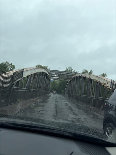 The image depicts a view from inside a vehicle driving across a wet, metal truss bridge under an overcast sky. The road surface is reflective, indicating recent rain. On the bridge, there is a single white car in the distance and part of a black SUV is visible on the right side of the frame. The bridge structure is made of rusted metal beams and supports, with metal fencing along the sides. Lush green trees and foliage are visible on either side of the bridge. The scene appears to be in a natural or semi-natural setting, with no visible buildings or urban infrastructure apart from the bridge itself and the vehicles. The image is taken from a low angle, looking forward through the windshield. While the exact location is not specified, the presence of a Truss bridge of this style might suggest it is an older, possibly heritage structure.