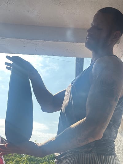 A man stands outdoors, balancing a dark, oblong object on his forearm. He is wearing a dark tank top with a camouflage pattern and striped pants. Tattoos are visible on his arms. The background shows a bright sky with some clouds and greenery. The image appears to be taken from a low angle, looking upwards. The overall lighting suggests it is daytime, possibly with direct sunlight casting shadows. No specific location in Heinebach, Germany is identifiable from the visual information present.