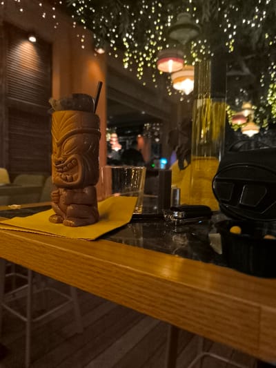 This photograph captures a dimly lit bar interior, likely in Palaio Faliro, Greece, showcasing a vibrant evening atmosphere. The central subject is a dark brown, intricately carved tiki mug, filled with a drink and garnished with a dark, possibly chocolate or fruit, element and a black straw. The mug rests on a bright yellow napkin positioned on a dark, marble-like counter surface.

To the right of the tiki mug, a clear glass sits next to a tall, slender bottle containing a yellowish liquid. Further right, a black case or bag prominently features the distinct "D" logo, indicative of the Diesel brand. Beside it, a small black container holds several round, yellow objects, resembling candies or nuts, suggesting bar snacks. A small metallic object, possibly a lighter, is also visible on the counter. The foreground shows a wooden bar top.

The background is softly blurred, revealing numerous small, warm fairy lights intertwined with foliage overhead, creating a sparkling, canopy-like effect. Several decorative hanging lamps with colorful shades emit a soft glow. Indistinct figures of people are visible in the distance, implying other patrons or staff, contributing to a lively yet relaxed social setting. The overall impression is one of an inviting and stylish establishment, perfect for an evening out.