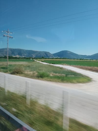 The image depicts a daytime, outdoor scene, likely captured from a moving vehicle, showing a landscape with agricultural fields and rolling hills under a clear blue sky. In the foreground, a blurred section of a paved road runs diagonally from bottom left to the center right, bordered by a blurred grassy area. To the left, a power pole with crossbars and electrical wires stretches upwards towards the sky. The middle ground features expansive green fields, some appearing to be cultivated crops, with a dirt path or unpaved road winding through them. In the background, a series of low, rounded hills or mountains form the horizon line, with some vegetation visible on their slopes. The overall impression is of a rural, agricultural area. The reference to "the city of Zarkos, Greece" is not directly verifiable from the visual information within the image itself but is provided as a contextual clue to the location.
