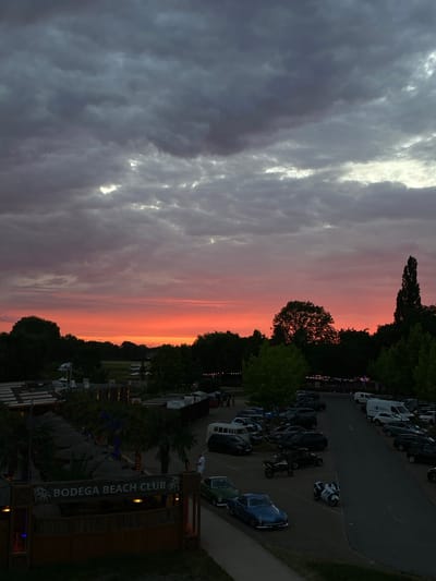 The image depicts an outdoor scene with a vibrant sunset creating a dramatic sky. In the foreground, a sign reads "BODEGA BEACH CLUB" with palm trees and thatched umbrellas. Adjacent to the club is a parking lot filled with various vehicles, including cars, vans, and motorcycles. Some people are visible near the club and in the parking lot, appearing to interact or move about. The background features a treeline and a field with what appear to be wind turbines in the distance. The scene suggests a gathering or event taking place at the beach club as dusk settles. The image is likely taken in the city of Rinteln, Germany, given the context provided.