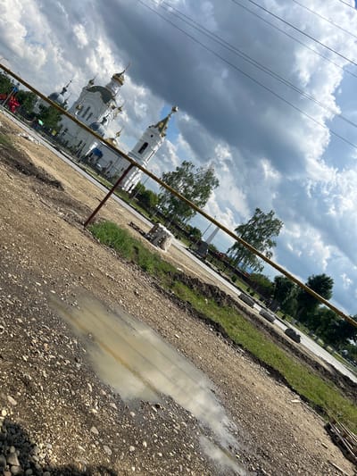 The media file depicts an outdoor scene, likely in Dondukovskaya, Russia, on a cloudy day. The dominant subjects are a white church with multiple golden domes and a construction site in the foreground. The church, with its elaborate architecture, suggests a place of worship. The construction site features a dirt and gravel path with several puddles, indicating recent rain. In the background, trees and a paved area with some building materials like pallets can be seen. The overall impression is one of ongoing development or renovation, juxtaposed with the established presence of the church. The lighting suggests it is daytime, but the heavy cloud cover casts a diffused light over the scene. There are no visible people clearly identifiable, and no text is legible within the frame. The tilted angle of the shot adds a dynamic element to the otherwise still scene.