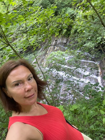In the city of Izmaylovka, Russia, a woman with short brown hair and fair skin is captured in a selfie. She is wearing a bright red, sleeveless top with a ribbed texture. Her expression is neutral, with her eyes looking directly at the camera. She is positioned in the foreground, with a lush green forest setting behind her. A cascading waterfall flows down a rocky incline in the background, surrounded by dense foliage. The overall scene depicts a natural outdoor environment.