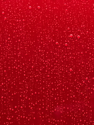 The image is a close-up, abstract view of a red, glossy surface with numerous water droplets of varying sizes. The droplets are scattered across the surface, creating a textured, almost bubbly appearance. The lighting suggests the surface is slightly reflective, as some droplets have highlights. There are no people, animals, buildings, or any discernible features of a specific location within the city of Восход Снт (фролово), Russia visible in the image. The focus is solely on the pattern and texture of the water droplets on the red surface.