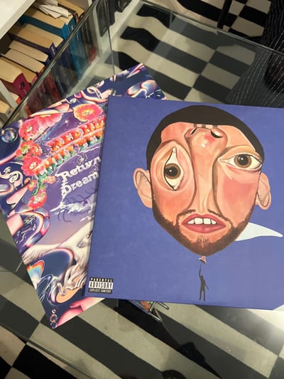Two vinyl records sit atop each other. The top record features an upside-down, surreal portrait of a person with exaggerated eyes and mouth, set against a solid blue background. A parental advisory sticker is visible in the bottom corner. The record beneath has a vibrant, colorful design with floral and dreamlike imagery, including bubbles and iridescent patterns. Both records are on a glass surface, reflecting the checkerboard pattern of a nearby surface, and a stack of books can be seen through the glass.