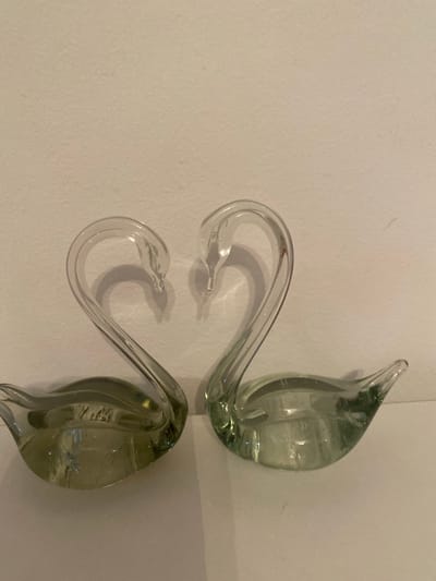 The image shows two clear glass swan figurines placed on a white surface against a neutral background. The swans are positioned facing each other, with their necks curved to form a heart shape. Their bodies are bulbous, and their heads are slightly pointed. The lighting creates shadows on the wall behind the figurines. There is no indication of any specific location within Жаңатұрмыс, Kazakhstan.
