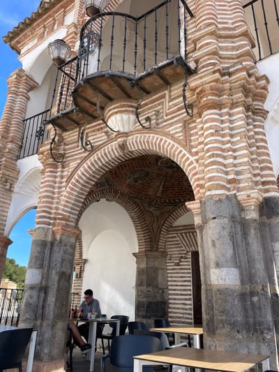 The image shows a low-angle view of the underside of an archway made of red and white brick. The archway is part of a larger building that is made of stone. The archway is located in Antequera, Spain (latitude: 37.014, longitude: -4.56) and leads into an open-air area. A person is sitting on a chair to the right of the archway, while a second person sits on another chair slightly to the left of the first person. There are additional chairs stacked near the first person and a table to their right. The scene is lit by the sun and is partially obscured by the architecture.