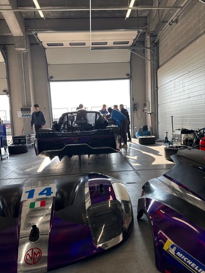 The image displays the interior of a garage bay at a motorsport facility in San Martino del Lago, Italy, likely the Cremona Circuit. A partially disassembled, purple and black prototype race car, stripped of its body panels, occupies the central background, elevated on stands. Its exposed engine bay and rear diffuser are clearly visible. Six to seven individuals are present: one young male in a dark hoodie stands on the left, observing, while a group of five to six men are gathered around the rear of the elevated race car, engaged in mechanical work or inspection. On the concrete floor in the foreground, two distinct race car body panels rest. The left panel is purple and black, featuring the number "14" in blue, an Italian flag decal, and an "NM" logo, alongside "EUROPEAN SERIES" text. The right panel, also purple and black, displays a yellow "MICHELIN" logo. The garage bay is brightly illuminated by natural light entering through a large, open overhead door at the rear, casting long shadows across the concrete floor. Overhead fluorescent lights are visible on the ceiling. An air compressor, various tires, and tools are distributed throughout the bay, alongside wall-mounted electrical panels and concrete support pillars.