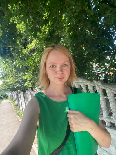 This is a vertical, selfie-style photograph taken outdoors, likely during the daytime on a sunny day. The subject is a light-skinned woman with blonde hair, wearing a green top. She is holding a green folder in her left arm, partially obscuring her torso. Her facial expression is neutral.
The background features lush green foliage from trees, suggesting a park or garden setting. To the right of the woman, a concrete or stone decorative balustrade is visible, and beyond that, a paved walkway or path stretches into the distance, lined with some greenery. The overall setting appears to be a public outdoor space in a somewhat urban or suburban environment, with hints of buildings in the background through the leaves.
No visible text is present in the image. The orientation of the photo is such that the top of the woman's head is towards the bottom right of the frame.