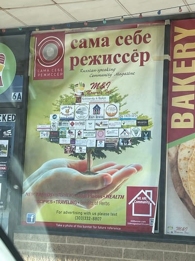 The image shows a large banner advertising "сама себе режиссер" (Sama Sebe Rezhisser), a Russian-speaking community magazine. The banner features an illustration of hands holding a tree with many small logos of businesses embedded within its branches. The background of the banner is green and yellow, with the magazine title prominently displayed in red Cyrillic and English. Below the tree, there are text elements listing "NEW TRENDS INTERVIEWS Cool Places HEALTH RECIPES TRAVELING Benefits of Herbs", and contact information for advertising. A Facebook logo and a QR code are also visible. The banner is displayed on the exterior of a building, likely a storefront, with a brick facade visible at the bottom. To the right of the banner, a sign for "BAKERY" is visible. The scene appears to be in a commercial area in Denver, Colorado, United States.