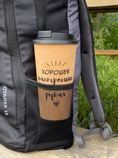 A close-up, eye-level shot shows a brown paper coffee cup with a black lid placed in the side mesh pocket of a black backpack. The cup is decorated with text that appears to be in Russian, reading "ХОРОШЕЕ НАСТРОЕНИЕ" (Good Mood) and a smaller inscription below that says "рубас" with a heart symbol. The backpack is black with grey mesh accents and straps. The background is blurred, showing green grass and a wooden surface. The image is of the exterior of a backpack with a coffee cup in its side pocket, with a natural outdoor setting in the background. The location of Petrovskoye, Russia is not ascertainable from the visual information presented in the image.