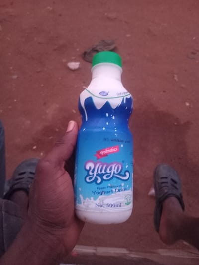 A person holds a bottle of Yugo Apple flavored Yogurt Drink in their hand. The bottle is blue and white, with a green cap, and reads "Net: 500ml". In the background is red soil, white debris, a pair of grey slip-on shoes, and what appears to be the person's jeans. The bottle label includes the word "Probiotics" in small print.