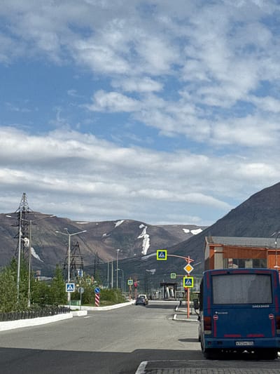 The image depicts a road in Norilsk, Russia, with a blue bus in the foreground. The bus is parked on the right side of the road, facing away from the viewer. It has the word "ЗАКАЗНОЙ" (meaning "custom-made" or "on demand" in Russian) written on its rear. The license plate of the bus is visible and reads "K925ME 124". A mountainous landscape with patches of snow is visible in the background, along with power lines and a building with an orange roof. Traffic signs, including pedestrian crossing signs and a traffic light, are present along the road. The sky is blue with scattered clouds. There are no people visible in the image.