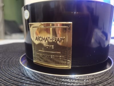 A close-up, eye-level photograph displays a dark, glossy cylindrical object, likely a candle or jar, positioned on a dark, woven circular placemat. A rectangular gold-colored label adhered to the object's front reads "AROMATHERAPY HOME PREMIUM EDITION." A smaller, oval, light-colored label, partially obscured, is visible on the upper left side of the object and appears to state "FERRERO ROCHER." The object rests on a circular silver-colored coaster featuring a patterned rim. The placemat exhibits a concentric circular weave pattern. The background is out of focus, revealing blurred reflections and indistinct room elements. No individuals are present. The scene is located in Velichkovo, Bulgaria.