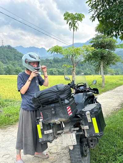 An outdoor, eye-level shot in Ngọc Đường Commune, Vietnam, captures a person beside a motorcycle on a dirt path, with expansive rice fields and mountains in the background under a cloudy sky.
The primary subject is an individual, likely a young adult, standing next to a dark-colored adventure motorcycle. The person is wearing a dark blue t-shirt, loose-fitting dark gray pants, and light gray sandals. They are in the process of adjusting a light gray full-face helmet with both hands, either putting it on or taking it off. Bracelets are visible on both wrists, and a tattoo can be seen on their lower left leg/ankle.
The motorcycle is equipped for touring, featuring hard-shell panniers (side cases) on both sides of the rear wheel. A black soft bag, possibly a backpack, is strapped to the back of the motorcycle, between the seat and the rear panniers. A yellow reflective strip is affixed to the right pannier, and red and white reflective strips are on the left. The motorcycle has spoked wheels, knobby tires, and a windscreen. A license plate is partially visible on the rear, displaying "29-A1 95" followed by additional numbers that are unclear.
The setting is a picturesque rural landscape. A narrow dirt or gravel path runs alongside a bright green and yellow rice paddy field, indicating the harvest season or stages of growth. Beyond the fields, lush green trees and distant mountains rise under an overcast sky, suggesting a bright but possibly humid day. Scattered power lines are visible in the upper background. The overall scene suggests a journey or stop along a scenic route.