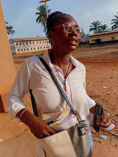 A woman with glasses and braided hair stands outside. She wears a white shirt and carries a two-toned purse. She holds a black smartphone. Palm trees are visible in the background, along with a tall, light-colored building with repeating windows. The ground appears to be reddish-brown soil. The lighting suggests a sunny day.