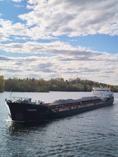 The image depicts a fully loaded cargo ship, named "ТOЛBУЯ" ("TOLBUYA"), traveling on a body of water, presumably a river or lake. The ship is dark-colored and appears to be carrying a large quantity of gravel or similar material. The water has moderate ripples. In the background, there is a shoreline with trees. The sky is blue with scattered white clouds. Based on the name of the ship and contextual information, the location is likely near Yakunina Gora, Russia.
