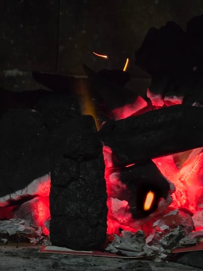 The image captures an active combustion scene located in Villa San Andrés, Argentina. The primary subjects are numerous dark, irregularly shaped pieces of charcoal, some of which are intensely glowing bright red, indicating high-temperature combustion as embers. Multiple small, flickering yellow-orange flames and bright orange sparks are visible emanating from the burning charcoal. Accumulations of white and light grey ash are present at the base of the charcoal pile, consistent with combustion byproducts. Underneath a portion of the ash, fragments of partially burned, possibly reddish paper or cardboard are discernible. The charcoal and ash rest on a dark, textured surface. The background is dark and indistinct, suggesting an enclosed fire containment structure. No individuals or animals are visible in the frame.