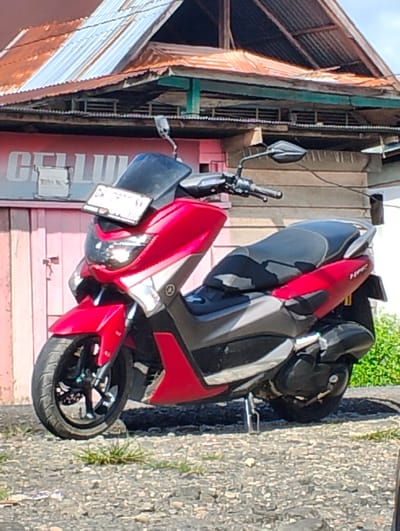 A red and gray Yamaha NMAX scooter is parked on a gravel surface in front of a building with a corrugated metal roof. The building appears to be a shop with a pink wooden door and a sign that reads "CELLULAR". The scooter is facing towards the viewer's left, and its license plate is visible. The background shows the building and some green foliage. The scene is outdoors during the day, with sunlight casting shadows.