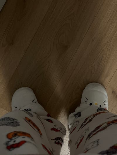 A pair of legs clad in white pajamas patterned with colorful drawings, possibly of dogs, stand on a wooden floor. Each foot is encased in a white, plush slipper, one of which features a Hello Kitty design on the toe. The floor is a light brown wood with grain patterns running diagonally across the frame, creating a simple yet cozy indoor scene.