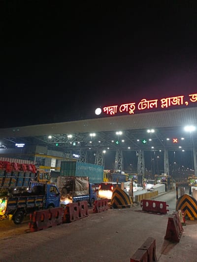 The image showcases the illuminated Padma Setu toll plaza at night. The dark sky contrasts with the bright signage above the toll booths. Numerous trucks are lined up in the foreground, waiting to pay their tolls. Striped barriers and red plastic blocks separate the lanes. Beyond the trucks, cars are visible passing through the booths, highlighted by their headlights. Overhead lights illuminate the entire scene, creating a stark and somewhat industrial aesthetic.