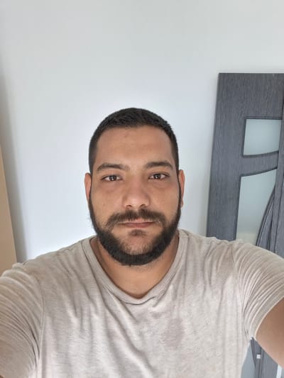 A medium shot, chest-up, selfie-style portrait of a man with dark hair and a beard. He is wearing a light-colored, short-sleeved t-shirt. The background is a plain, off-white wall with a gray door visible to the right of the man. The man's head is slightly tilted, and he is looking directly at the camera with a neutral expression. The lighting is soft and even. There are no discernible features that would indicate the location within the city of Dolna Malina, Bulgaria.