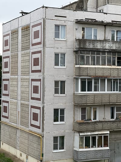 This is an eye-level, medium shot of a tiled, multi-story apartment building in Rzhev, Russia. The building has a grey, tiled facade with a pattern of brown squares within white squares on one section. There are multiple windows and balconies on the building, some of which are enclosed. The balconies have concrete decorative elements. Several birds are perched on the roof and one is in flight. A yellow gas pipe runs along the side of the building. The sky is overcast.