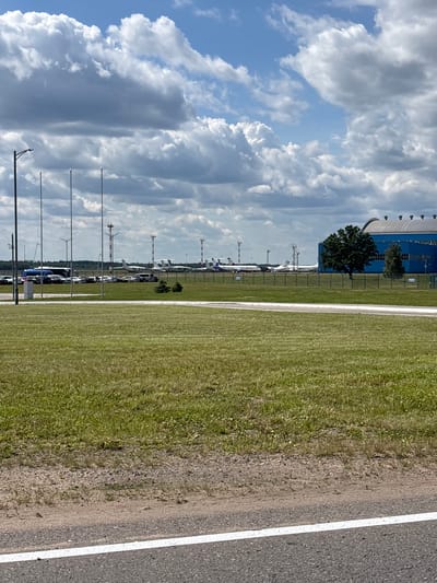 The media file depicts an outdoor scene at an airfield. In the foreground, a paved surface with a white line marking is visible, transitioning to a grassy area. Beyond the grass, a line of parked cars is observed. Further in the background, several airplanes are parked on the tarmac, with some appearing to be small private aircraft. A large, blue industrial building with a curved roof and rows of windows is situated behind the parked planes and trees. The sky is filled with dramatic, large cumulus clouds, suggesting either a partly cloudy day with potential for weather changes, or a recent storm. There are no people visible in the image. The lighting suggests it is daytime. The scene conveys a sense of activity at an airfield, likely a small to medium-sized airport or private airstrip, given the types of aircraft and the presence of a large hangar. The location context is Пекалінскі сельскі Савет, Belarus.