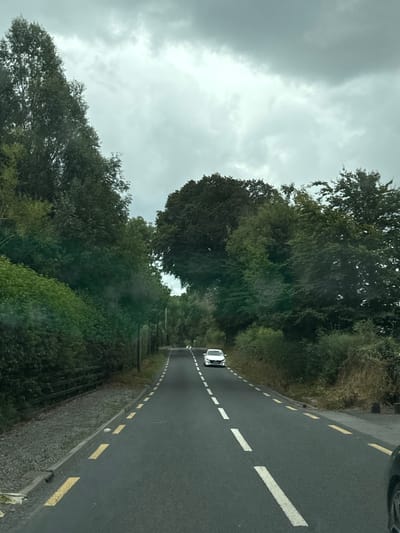 The image depicts a view from a vehicle driving on Eyrefield Road in Ireland. The road is asphalt with solid yellow lines marking the edge and broken white lines dividing the lanes. Dense green trees and foliage line both sides of the road, with a wooden fence visible on the left. The sky is overcast and cloudy. A white car is visible ahead, traveling in the same direction. The overall impression is of a rural or semi-rural setting on a cloudy day.