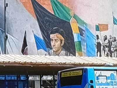 This media file depicts a mural on a building. The mural features a central figure, a young man with dark hair and a serious expression, wearing a headband and a checkered shirt. He is surrounded by various flags in vibrant colors such as black, blue, green, and orange. To the right of the central figure, silhouettes of people are visible, suggesting a crowd or procession. The mural appears to be painted on a textured surface and is positioned above a corrugated metal roof. Below the mural, a blue and white bus is partially visible, with some text in Arabic script on its side. The scene suggests a commemorative or historical event, possibly a protest or a celebration, given the presence of flags and figures. The lighting suggests it is daytime, with no specific weather conditions evident. The location context provided is Malmö, Sweden, but there are no direct visual cues within the image to confirm this. The visible text on the bus appears to be an advertisement or route information.