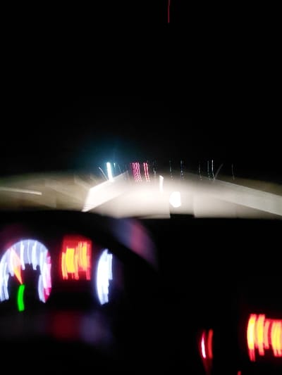 Blurred and abstracted lights dominate a black background. The right side features stacked, hazy red lights and a section with glowing numbers and a speedometer with a green indicator. A bright, vertical white streak extends from the top center, blurring into a column of light with embedded, colorful streaks. The overall image is dark and difficult to interpret.