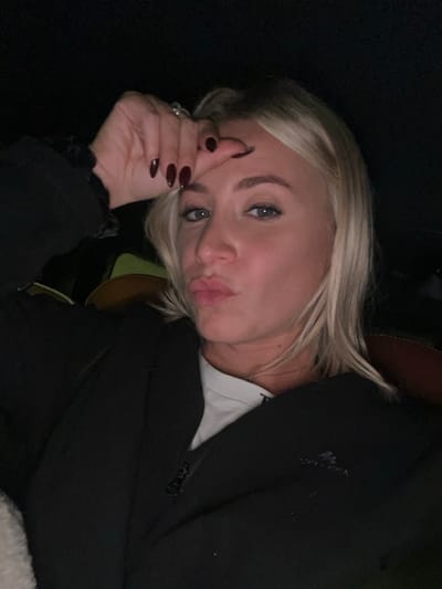 A fair-skinned woman with blonde hair is looking at the camera with pursed lips. Her left hand is near her forehead. She has dark polished nails and a ring on one finger. She is wearing a black blazer over a white t-shirt. The background is dark, with a glimpse of a cushioned surface beneath her. The image is rotated, presenting a sideways view.