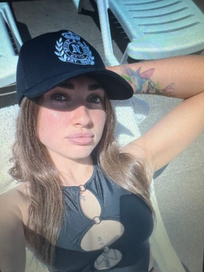 The image shows a woman wearing a black baseball cap with a crest on it and a dark, possibly black, sleeveless top with circular cutouts. She has long, light brown hair and is looking towards the camera with a neutral expression. Her arm is resting on something behind her head, and a colorful tattoo is visible on her upper arm. The background appears to be outdoors, with a blurry view of white lounge chairs and a textured surface that could be a patio or ground. There is a noticeable shadow cast across the woman's face from her cap. The image quality is low, with visible pixelation and compression artifacts. There is no indication of the location being Protasovo, Russia.
